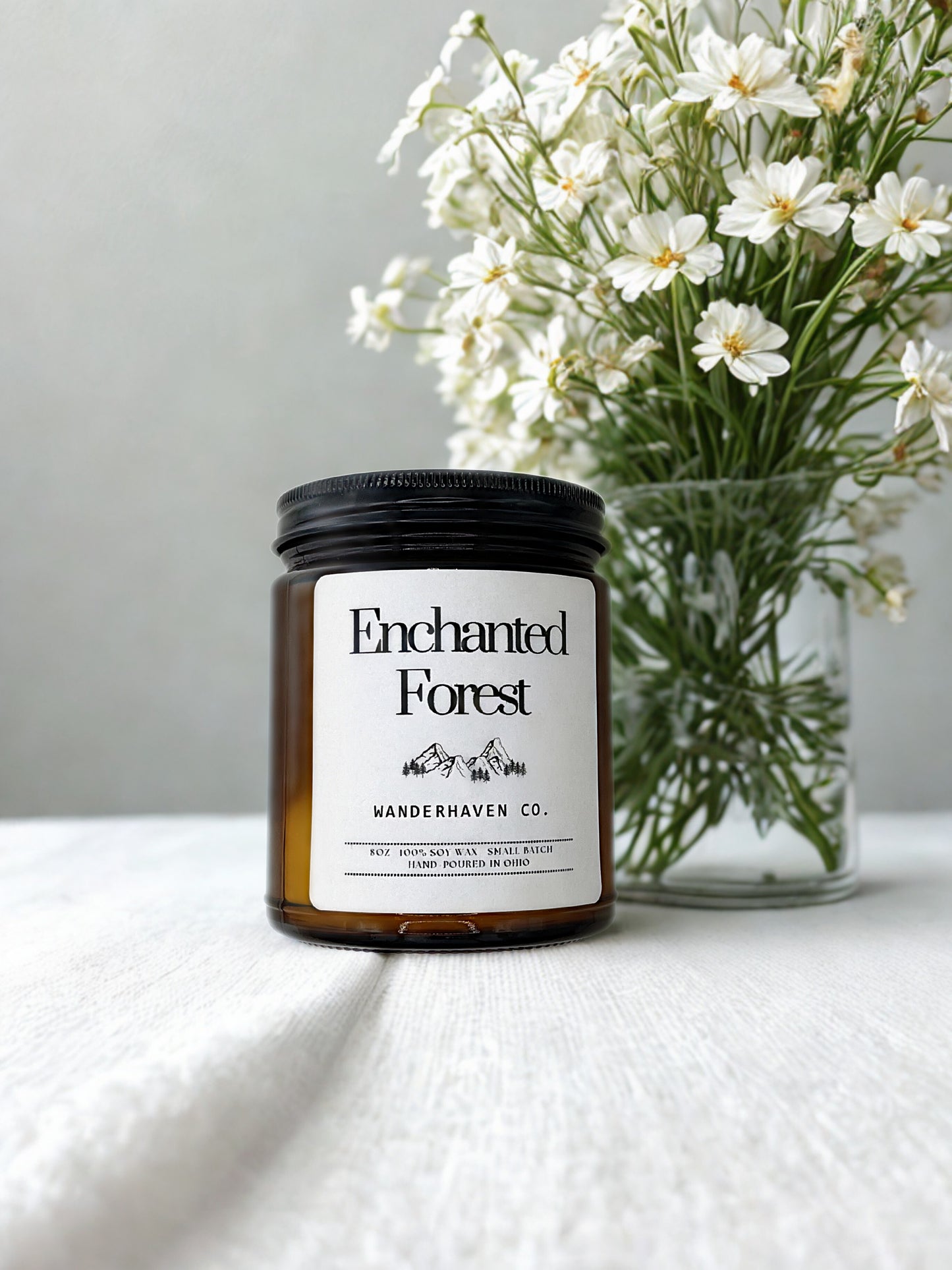 Candle labeled 'Enchanted Forest' by Wanderhaven Co. with a vase of white flowers in the background.
