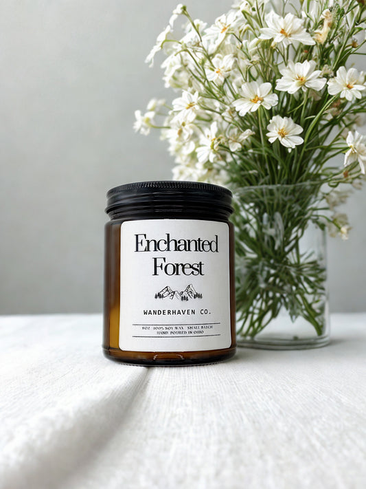 Candle labeled 'Enchanted Forest' by Wanderhaven Co. with a vase of white flowers in the background.