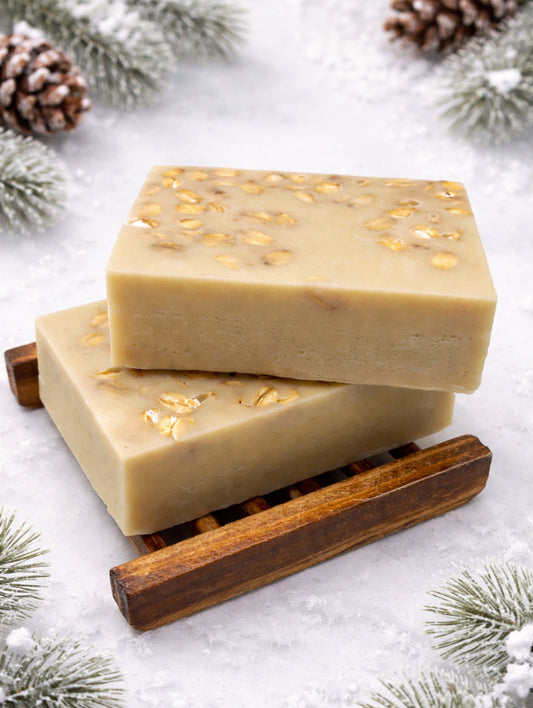 Two bars of soap on a wooden soap dish with pine cones and branches in the background